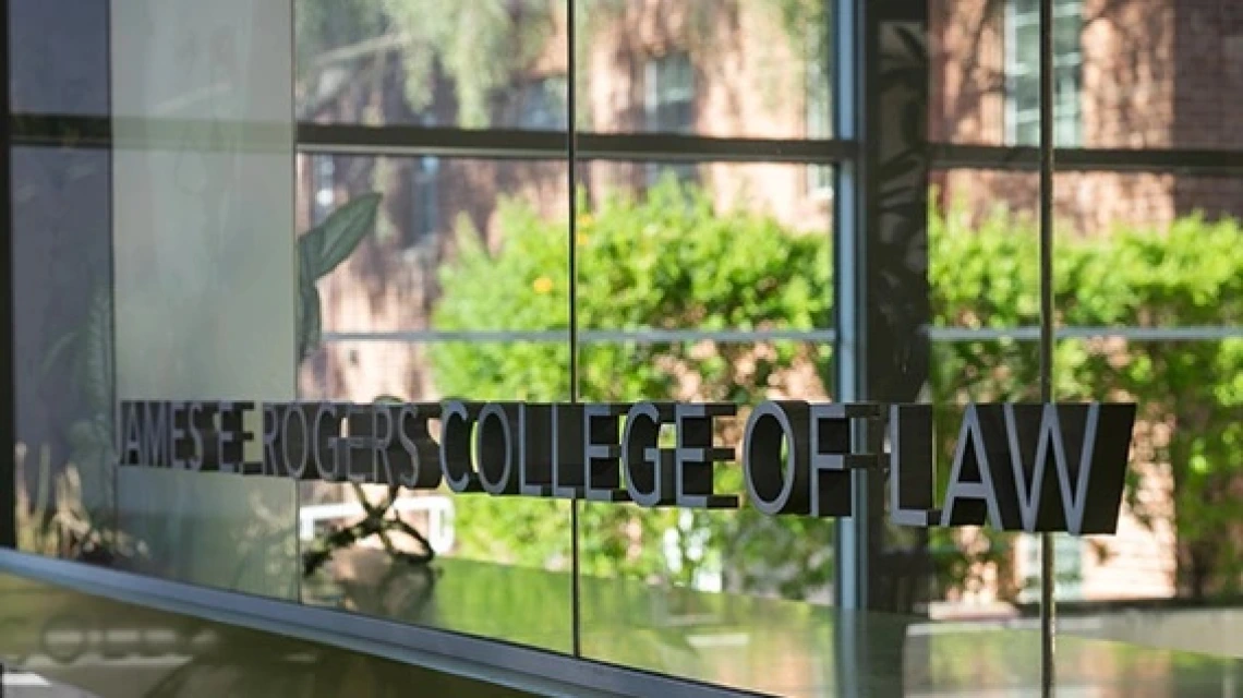 Law School windows reflect greenery and brick building behind 'James E. Rogers College of Law' sign