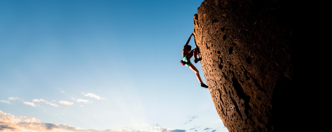 A person climbs the face of a large rock while the sun beams behind clouds in a blue sky behind them